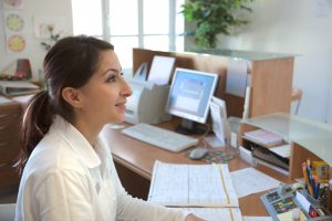 A young woman behind a GP receptionist desk, smiling and looking helpful.